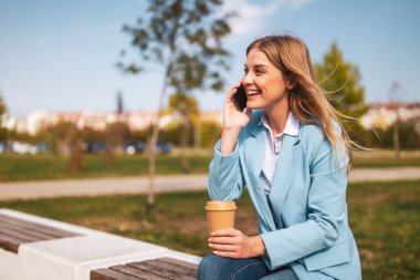 Gülümseyen iş kadını telefonda konuşuyor ve bankta otururken kahve içiyor..