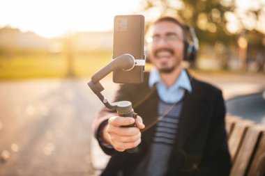 usinessman vlogger açık havada otururken ve güneşli bir sonbahar öğleden sonrasında vlog kaydederken cep telefonu kullanıyor. Kulaklık ve gözlük takıyor. Telefona ve telsize odaklan..