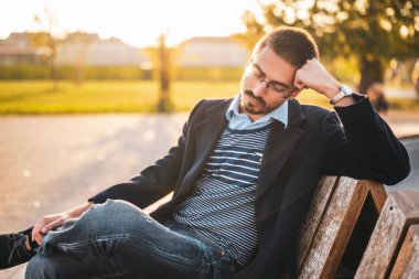 Güneş batarken parktaki bankta oturan gözlüklü üzgün iş adamı.