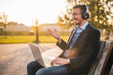 Gözlüklü ve kulaklıklı gülümseyen işadamı güneşli bir sonbahar öğleden sonrasında parkta otururken dizüstü bilgisayar kullanıyor ve konuşuyor..
