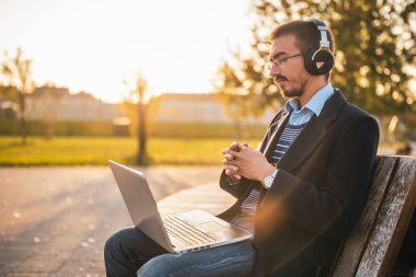 Gözlüklü ve kulaklıklı bir işadamı güneşli bir sonbahar öğleden sonrasında parkta otururken dizüstü bilgisayar kullanıyor ve çalışıyor..