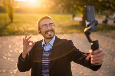 Businessman vlogger in coat wearing in-ear headphones and eyeglasses showing ok sign while using mobile phone with gimbal. He is standing in the park and recording vlog on a sunny autumn afternoon. clipart