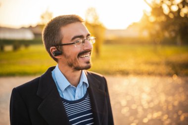 Parkta güneşli bir sonbahar öğleden sonrasında kürklü, gözlüklü ve kulaklık takan gülümseyen iş adamının portresini kapat..