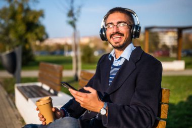 Gözlüklü mutlu işadamı dışarıda kahve içerken kulaklık ve cep telefonu kullanıyor. Güneşli bir sonbahar gününde parkta bankta oturuyor ve dinleniyor..