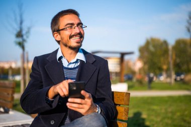 Parkta güneşli bir sonbahar gününde bankta otururken gözlüklü mutlu bir işadamı cep telefonu kullanıyor..