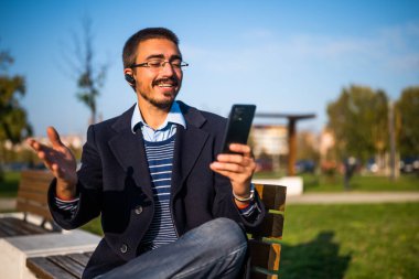 Parkta güneşli bir sonbahar gününde bankta otururken akıllı telefon ve kulaklık takan gözlüklü gülen bir işadamı..