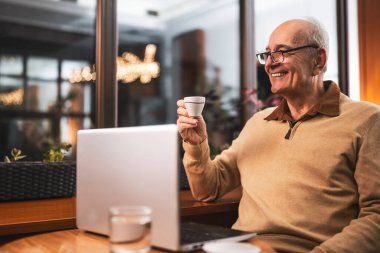 Gözlüklü, gözlüklü, gülümseyen, kahve içen ve rahat bir kafede dizüstü bilgisayar kullanan son sınıf öğrencisi..
