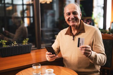 Rahat bir kafede kahve içerken akıllı telefon ve kredi kartı kullanan rahat giyinen mutlu kıdemli bir adam..