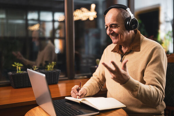 Cheerful senior man in casual clothing using wireless headphones, laptop and taking notes while attending online class. He is talking and gesturing during a video call on his computer in the cafe.