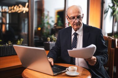 Kendine güvenen, şık takım elbiseli, gözlüklü, dizüstü bilgisayarda çalışan ve kahve içip kafede otururken belgeleri inceleyen kıdemli iş adamı..