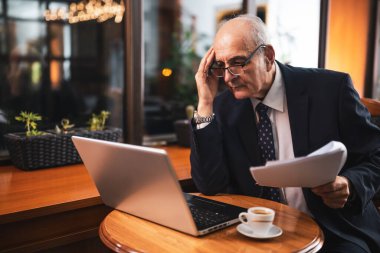 Takım elbiseli, gözlüklü, dizüstü bilgisayarda çalışan ve kahve içip şehir kafesinde otururken belgeleri inceleyen stresli kıdemli işadamı..