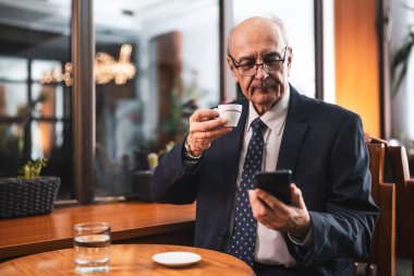 Şık takım elbiseli, kendine güvenen kıdemli işadamı cep telefonu kullanıyor ve rahat bir kafede oturup kahve içiyor..
