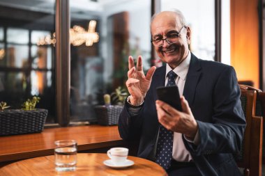Şık gözlüklü başarılı kıdemli işadamı cep telefonu kullanıyor ve rahat bir kafede oturup kahve içiyor..