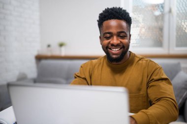 Keyifli Afro-Amerikalı adam rahat evinde masada otururken dizüstü bilgisayarla çalışıyor..
