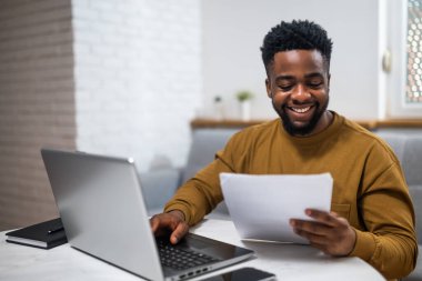 Gülümseyen genç Afrikalı işadamı evinde çalışırken bilgisayarını kullanıyor ve belgeleri inceliyor..