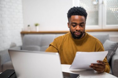 Kendine güvenen Afro-Amerikan bir adam evinde çalışırken bilgisayarını kullanıyor ve belgeleri inceliyor..