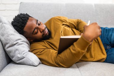 Rahat Afro-Amerikalı bir adam evinde kanepede dinlenirken kitap okumaktan zevk alıyor..