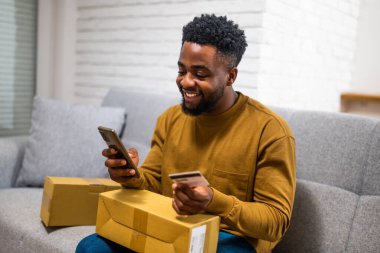 Neşeli genç Afro-Amerikalı adam teslimat kutusunu tutuyor, cep telefonu ve kredi kartını evinde rahat bir şekilde kanepede otururken cep telefonu ve kredi kartı olarak kullanıyor..