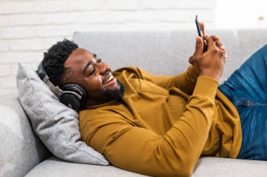 Kablosuz kulaklık ve cep telefonu kullanan neşeli Afro-Amerikalı adam rahat bir şekilde oturma odasındaki kanepede uzanıyor..