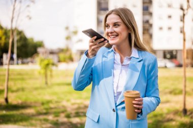 Genç iş kadını hareket halinde, akıllı telefonla konuşuyor ve parkta kahvesini yudumluyor..
