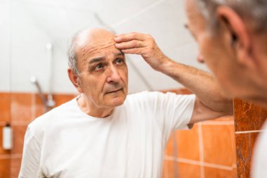 Endişeli kıdemli adam banyoda dikilirken derisini ve kırışıklıklarını analiz ediyor..