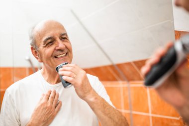 Sakalını tıraş etmek için tıraş makinesi kullanırken banyo aynasına bakan mutlu kıdemli adam..