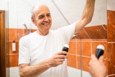 Kendinden emin kıdemli bir adam günlük bakım rutininin bir parçası olarak banyo aynasının önüne deodorant sürüyor..