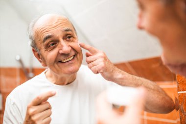 Yüzüne nemlendirici sürerken banyo aynasında yansımasına bakan neşeli yaşlı adamın portresi. 