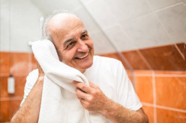 Neşeli son sınıf öğrencisi yüzünü banyoda beyaz bir havluyla kurutuyor aynadaki yansımasına gülümsüyor..