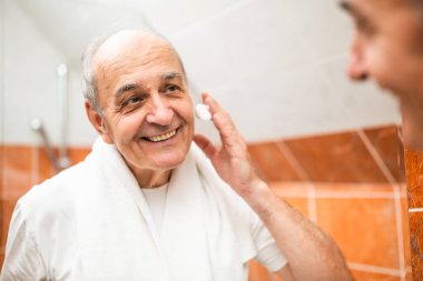 Boynunda havluyla banyodaki aynadaki yansımasına bakarken tıraş kremi süren kendinden emin yaşlı bir adamın portresi.. 