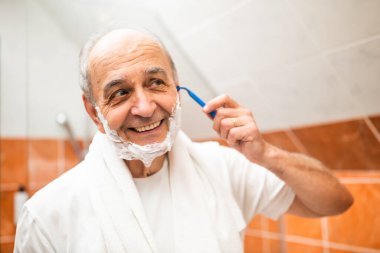 Banyoda aynanın önünde dikilirken tıraş olurken sakalını jiletle tıraş etmekten hoşlanan mutlu kıdemli adam..