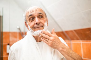 Tıraş kremi sürerken banyodaki aynaya bakan yaşlı adam..