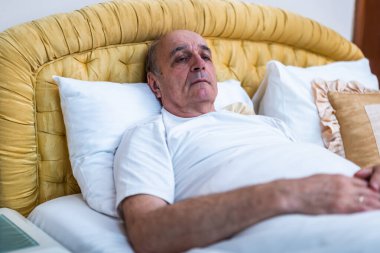Kıdemli adam sabahın erken saatlerinde yatakta uzanıyor, derin üzüntü ve zihinsel yorgunluğunu dile getiriyor. Yorgunluğun, depresyonun ve yaşlanmanın duygusal bedelinin güçlü bir tasviri..