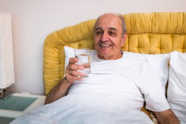 Neşeli son sınıf öğrencisi yatağında uyandıktan sonra su içmekten zevk alıyor. Emeklilikte sağlıklı yaşam tarzı ve sabah rutininin sembolü.