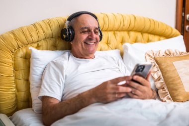 Yatak odasında rahat bir şekilde otururken pijamalı, kablosuz kulaklık ve cep telefonu kullanan neşeli bir son sınıf öğrencisi..