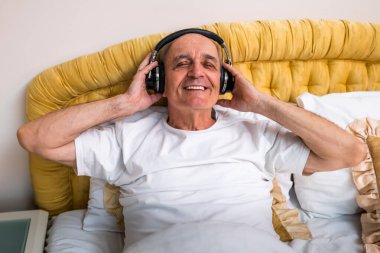 Gülümseyen son sınıf öğrencisi kablosuz kulaklıkla müzik dinlemekten ve yatakta dinlenmekten zevk alıyor. Onun mutlu ifadesi emeklilikteki rahatlığı ve memnuniyeti vurguluyor.