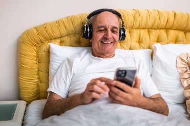 Kulaklıklı, pijamalı neşeli bir son sınıf öğrencisi evinde dinlenirken akıllı telefon kullanıyor..