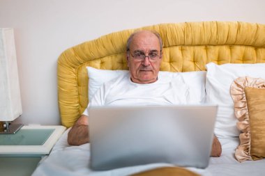 Gözlüklü, pijamalı, dizüstü bilgisayarı olan ve sabah kalktıktan sonra yatakta oturan son sınıf öğrencisi..