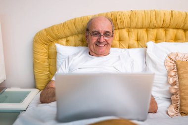 Gözlüklü, pijamalı, evinde rahat bir yatakta otururken laptopta çalışmaktan zevk alan neşeli son sınıf öğrencisi..