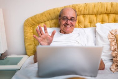 Gözlüklü, sabahları pijama giymiş, dizüstü bilgisayarındaki bir video görüşmesi sırasında konuşurken ve el kol hareketi yaparken mutlu bir son sınıf öğrencisi..