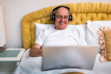 Pijamalı, kablosuz kulaklık takan ve rahat yatak odasında yatağında laptopla çalışan mutlu yaşlı bir adam..