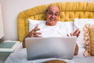 Yatakta otururken dizüstü bilgisayar kullanan pijamalı neşeli bir son sınıf öğrencisi. Konuşuyor ve gülümsüyor. Evinin rahatlığıyla konuşuyor.. 