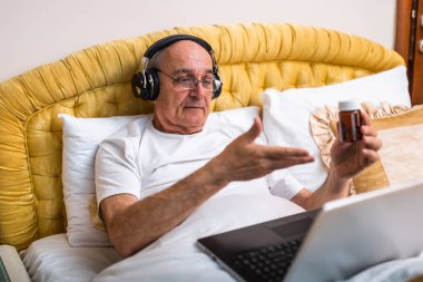 Kulaklıklı yaşlı adam ilaç şişesi tutuyor ve yatağında yatarken laptopta doktorla konuşuyor. Evinden gelen görüntülü arama ile tıbbi danışmanlık alıyor..