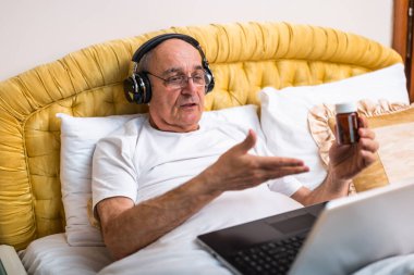 Kablosuz kulaklıklı yaşlı adam yatakta oturuyor ve bilgisayarından sağlık hizmeti uzmanıyla iletişim kuruyor. Tıbbi durumunu ve tedavi seçeneklerini tartışıyor..