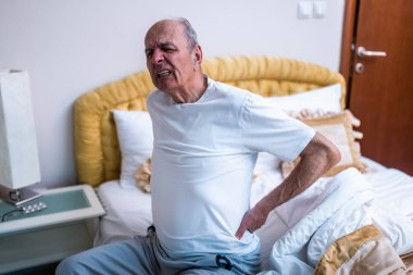 Yaşlı adam uyandıktan sonra sırt ağrısı çekiyor ve sabahın erken saatlerinde yatakta oturuyor..
