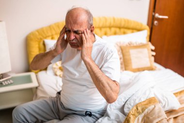 Sabah kalktıktan sonra baş ağrısıyla boğuşan ve hafifçe aydınlatılmış yatak odasında, yatakta oturan,.