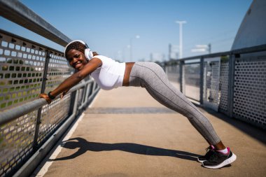 Kulaklık takan, şınav çeken ve şehir köprüsünde egzersiz yapan kararlı, Afro-Amerikan bir kadın. Gücünü, kas gücünü ve sağlıklı yaşam tarzına bağlılığını gösteriyor..