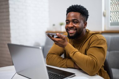 Genç Afro-Amerikalı adam dizüstü bilgisayar kullanırken gülümsüyor ve oturma odasında telefonla konuşuyor, uzaktan çalışıyor..