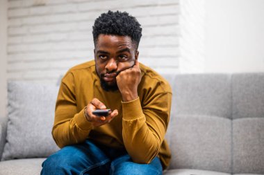 Genç Afrikalı adam evinde kanepede otururken sıkılıyor ve televizyon kanallarını değiştiriyor..