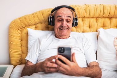 Gülümseyen yaşlı adam rahat yatak odasında cep telefonu ve kablosuz kulaklık kullanıyor..